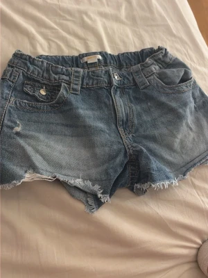 Blå jeansshorts med fransig kant - Säljer ett par klassiska blå jeansshorts med fransig kant och slitna detaljer. Shortsen har normal passform, fickor både fram och bak samt knapp och dragkedja framtill. Perfekta för sommaren och ger en avslappnad look.