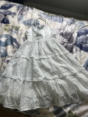 Ljusblå blommig långklänning - Söt långklänning i ljusblått och vitt med småblommigt mönster. Klänningen har smock i midjan, volangdetaljer och tunna axelband som knyts i nacken. Perfekt för varma dagar och ger en romantisk vibe.