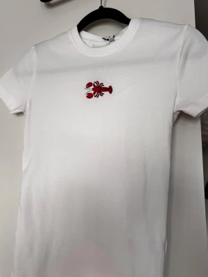 Vit t-shirt med röd kräfta - Snygg vit t-shirt med en broderad röd kräfta på bröstet. T-shirten har rund hals och korta ärmar. Materialet är supermjukt som passar perfekt till en avslappnad stil. Helt oanvänd, tog med den ut ur affären och hann aldrig prova och kände inte att den satt superbra när jag kom hem, sen hann jag aldrig lämna tillbaka🥲Synd för den är supersöt!