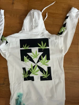 Vit hoodie med grafiskt tryck och huva - Säljer en vit hoodie från Off-White med stor grafisk svart logga och gröna blad på ryggen och ärmarna. Hoodien har huva med dragsko och ribbade muddar. Materialet är mjukt och bekvämt, perfekt för en streetwear-look.