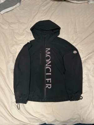 Svart vindjacka från Moncler - Snygg svart vindjacka från Moncler med stor logga vertikalt på framsidan och klassisk Moncler-patch på ärmen. Jackan har huva, dragkedja framtill och justerbara muddar. Perfekt för blåsiga dagar och streetwear-stil.