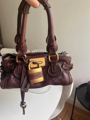 Chloe Paddington väska  - Säljer en mörkbrun Chloe Paddington handväska i läder med guldfärgade metalldetaljer och ett stort dekorativt hänglås framtill. Väskan har dubbla handtag, nitar och sömmar som ger en cool vintagekänsla. Perfekt accessoar för dig som gillar statementväskor. 28 cm bred och 12 hög