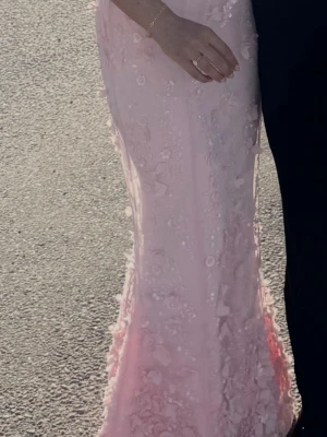 Ljusrosa långklänning med spets - Elegant ljusrosa långklänning med vackra spetsdetaljer och broderade blommor över hela klänningen. Figurnära passform som framhäver siluetten. Perfekt för speciella tillfällen där du vill känna dig extra fin.