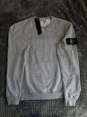 Grå sweatshirt från Stone Island - Snygg grå sweatshirt från Stone Island med klassisk rund hals och ribbade muddar. Tröjan har den ikoniska Stone Island-loggan på vänster ärm och är tillverkad i mjukt bomullsmaterial. Perfekt för en clean och stilren look.
