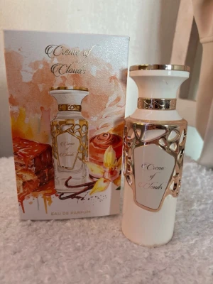 Crème of Clouds Eau de Parfum - Elegant parfymflaska i vitt med guldfärgade detaljer och dekorativt mönster.100ml Flaskan har en exklusiv look och levereras i en snygg ask med motiv av vanilj och karamell. Doften heter Crème of Clouds och ger en lyxig och söt känsla.