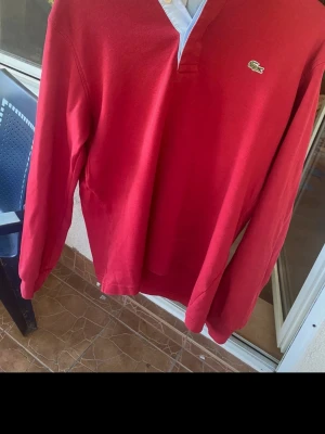 Röd långärmad tröja från Lacoste - Snygg röd långärmad tröja från Lacoste med klassisk krokodillogga på bröstet. Tröjan har en v-ringad krage med vit insida och är tillverkad i mjukt bomullsmaterial. Perfekt för en stilren och avslappnad look.