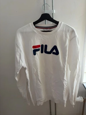 Vit sweatshirt från FILA - Vit sweatshirt från FILA med klassisk logga i rött och blått framtill. Tröjan har rund halsringning och långa ärmar. Perfekt för en sportig och avslappnad stil.