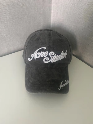 Acne Studios Keps - Säljer en grå keps från Acne Studios med broderad vit logga framtill och på skärmen. Kepsen har klassisk böjd skärm och justerbar rem bak. Tillverkad i tvättad bomull för en avslappnad look.