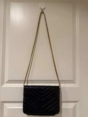 Svart quiltad axelväska med kedja - Snygg svart axelväska i quiltat mönster med guldfärgad kedjerem. Väskan har ett kuvertformat lock med magnetknäppning och insida i svart material. Perfekt för dig som gillar stilrena accessoarer med en lyxig touch.