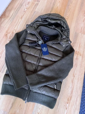 Leon Marcel hybridjacka med avtagbar huva – storlek M - Stilren Leon Marcel-jacka i mörk olivgrön/brun färg. Jackan har vadderad fram- och baksida, stickade ärmar, ribbad krage och avtagbar huva. Försedd med dragkedja framtill samt praktiska sidofickor med dragkedja.  Storlek: M Skick: 9,5/10 – mycket fint skick Elegant och varm modell som passar perfekt till höst, vinter och vår. Säljs enligt bilderna.