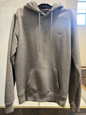 Grå hoodie från Gant - Snygg grå hoodie från Gant med broderad logga på bröstet. Klassisk modell med huva, dragsnören och stor magficka framtill. Perfekt för en avslappnad och stilren look.