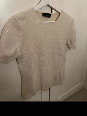 Beige stickad topp med paljetter - Snygg beige stickad topp med korta ärmar och rund hals. Toppen har små diskreta paljetter som ger ett subtilt skimmer och ett ribbat mönster. Perfekt för dig som vill ha något enkelt men ändå lite extra.
