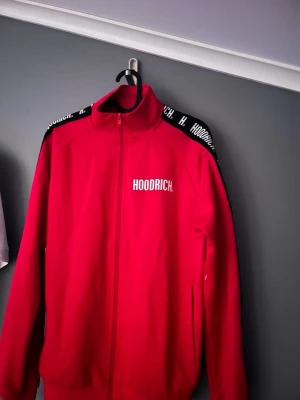 Röd zip hoodie från Hoodrich - Snygg röd hoodie från Hoodrich med dragkedja framtill och vit logga på bröstet. Svarta detaljer med vit text längs axlarna ger en sportig vibe. Tillverkad i mjukt material och har hög krage för extra stilpoäng.