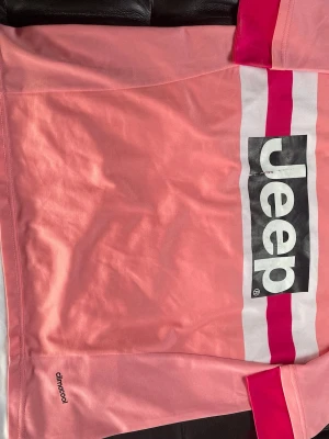 Juventus rosa fotbollströja POGBA - Juventus fotbollströja i rosa med svarta och vita detaljer samt Jeep-logga på framsidan. Tröjan har klubbmärke med tre stjärnor broderat på bröstet och är tillverkad i lätt och ventilerande climacool-material. Perfekt för fotbollsträning eller som supporterplagg.