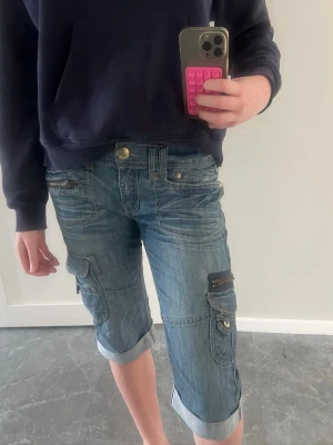 Blå cargoshorts i denim - Säljer ett par blå cargoshorts i denim med flera fickor och dragkedjedetaljer. Shortsen har uppvikta benslut och sitter midwaist. Perfekta för en avslappnad och cool stil.