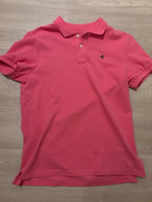 Rosa pikétröja med krage - Säljer en rosa pikétröja från La Martina med klassisk krage och två knappar framtill. Tröjan har korta ärmar och en liten broderad logga på bröstet. Perfekt för en avslappnad och stilren look.