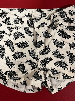 Vita shorts med svart bladprint från H&M - Säljer ett par vita shorts från H&M med svart bladmönster. De har klassisk passform, fickor fram och bak samt hällor för bälte. Perfekta för sommarens alla äventyr och är gjorda i bomull för en skön känsla.