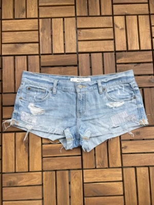 Ljusblå denimshorts med slitningar - Säljer ett par ljusblå denimshorts från Abercrombie & Fitch med råa kanter, slitningar och patchade detaljer. Klassisk femficksmodell med låg midja och avslappnad passform. Perfekta för varma dagar och en cool streetstyle-look. Midjemått 44cm