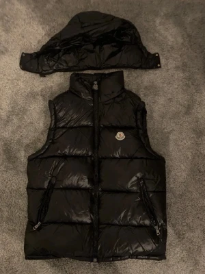 Svart dunväst från Moncler - Snygg svart dunväst från Moncler med avtagbar huva och två dragkedjeförsedda fickor. Västen har ett klassiskt Moncler-emblem på bröstet och är tillverkad i glansigt material med quiltad design. Perfekt för lager-på-lager och streetstyle.