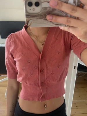 Kort rosa kofta från KappAhl - Snygg kort kofta i rosa från KappAhl med v-ringning och knappar framtill. Koftan har korta puffärmar och ett diskret mönster i stickningen. Perfekt att matcha med höga jeans eller kjol. Materialet är mjukt och stretchigt.