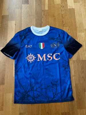 Napoli fotbollströja blå EA7 - Snygg blå Napoli fotbollströja från EA7 med vita och svarta detaljer. Tröjan har klubbmärke, italienska mästerskapsmärket och MSC-logga på bröstet. Rund halsringning i vitt och diskret mönster över hela tröjan.