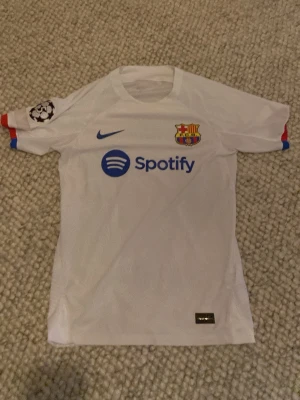 Vit FC Barcelona matchtröja Nike - Snygg vit FC Barcelona matchtröja från Nike med klubbmärke, blå Nike-logga och Spotify som sponsor framtill. Champions League-märke på ärmen och färgdetaljer i rött och blått vid ärmslut. Tillverkad i funktionsmaterial, perfekt för fotbollsträning eller match.
