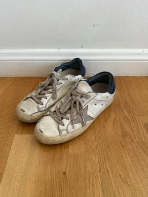 Golden Goose vita sneakers med stjärna - Snygga vita sneakers från Golden Goose med grå stjärndetalj på sidan och mörkblå hälkappa. Skorna har grå snörning och klassisk låg modell. Tillverkade i läder med slitna detaljer för en cool, avslappnad look.