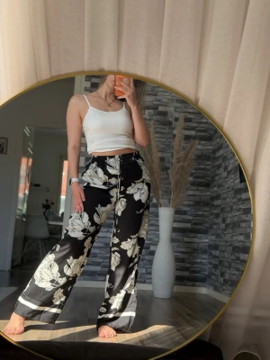 Svart blommigt loungewear-set - Snyggt loungewear-set med svart bas och stora vita blommor. Setet består av vida byxor med dragsko i midjan och matchande skjorta med långa ärmar och knappar. Perfekt för chill dagar hemma. Materialet ser ut att vara mjukt och följsamt.