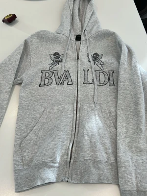 Grå BVALDI hoodie med strassmotiv - Snygg grå hoodie från BVALDI med glittrande strassdetaljer i form av text och änglar på framsidan. Hoodien har dragsko vid huvan och är tillverkad i mjukt material som känns skönt mot huden. Perfekt för dig som gillar unika detaljer och streetstyle.