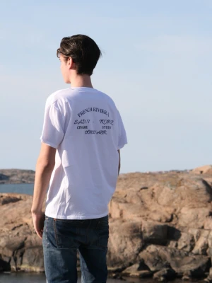 Vit t-shirt från Civaré( French Riviera Collection) - Vit t-shirt från Civaré med blå texttryck på både fram- och baksida. På ryggen står det 'French Riviera', 'Saint-Tropez' och 'Côte d'Azur' i snygg font. Klassisk passform och rund hals. Perfekt för dig som gillar stilrena plagg med en twist.