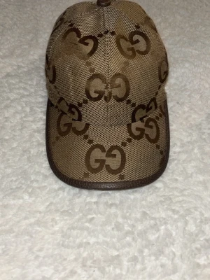 Beige keps med GG-mönster från Gucci - Snygg beige keps från Gucci med det klassiska GG-monogrammet i brunt över hela kepsen. Kepsen har böjd skärm och är tillverkad i ett tåligt tygmaterial med bruna detaljer längs kanten. Perfekt för dig som vill ha en exklusiv och trendig accessoar.