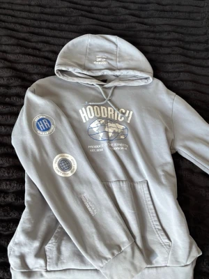 Grå hoodie från Hoodrich  - Snygg grå hoodie från Hoodrich med stor huva och vita tryck på bröstet och ärmen. Framtill finns en stor ficka och coola detaljer som texten 'PRODUCT OF THE STREETS' och globgrafik. Materialet är mjukt bomullstyg, perfekt för en avslappnad streetwear-look.