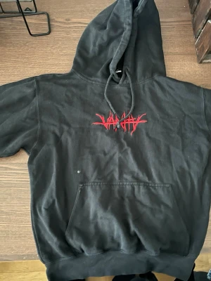 No vanity Juice Wrld merch - Säljer en svart hoodie med huva och känguruficka framtill. På bröstet finns en röd, grafisk logga i streetwear-stil. Hoodien har ribbade muddar vid ärmar och nederkant samt dragsko i huvan. Perfekt för dig som gillar en edgy och avslappnad look.