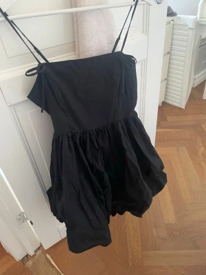 Svart kort klänning med tunna band - Säljer en svart kort klänning med tunna axelband och voluminös kjol. Klänningen har en enkel, stilren design med knyt vid axlarna och en luftig passform. Perfekt för dig som gillar minimalistisk stil.