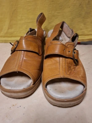 Bruna kilklackade sandaler i läder - Säljer ett par bruna sandaler från Wörishoff med bredd kilklack i kork och justerbara remmar med metallspänne. Sandalerna har öppen tå och häl, och är tillverkade i läder med ljus innersula. Perfekta för dig som vill ha både stil och komfort.