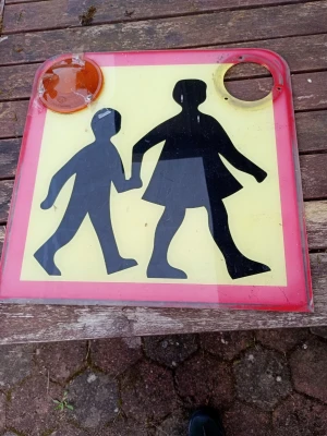 Skyltskylt med barnmotiv - Unik fyrkantig skylt i plast med svart siluett av två barn som håller varandra i handen på gul bakgrund och röd ram. Skylten har en orange reflex i ena hörnet och ett runt hål i det andra. Perfekt som dekoration eller samlarobjekt.. fattas ett rent plast skydd.