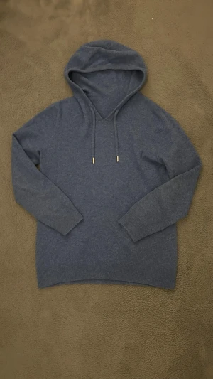 Merinoull hoodie - En stilren mörkgrå hoodie med huva och dragsnören med metalländar. Tillverkad i ett mjukt, stickat material som känns skönt mot huden. Långa ärmar med ribbade muddar och enkel design utan tryck eller logga. Perfekt för en avslappnad look.