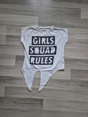 Vit t-shirt med tryck och asymmetrisk nederkant - Vit t-shirt från Firefly med svart texttryck 'GIRLS SQUAD RULES' på framsidan. T-shirten har korta ärmar och en unik asymmetrisk, spetsig nederkant framtill. Perfekt för dig som vill sticka ut med en edgy och cool stil. Den har några små hål, se bilder.