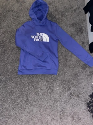 Blå hoodie från The North Face - Säljer en blå hoodie från The North Face med stor vit logga på bröstet. Hoodien har luva och magficka samt ribbade muddar vid ärmslut och nederkant. Perfekt för dig som vill ha en snygg och bekväm tröja med streetwear-vibe. Den är väldigt skön att ha på sig och passar med nästan allt
