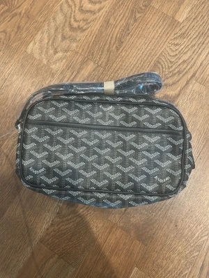 Svart axelväska  - Snygg svart axelväska med klassiskt mönster i vitt och grått. Väskan har en rektangulär form och justerbar axelrem. Materialet är canvas med detaljer i läder. Perfekt för dig som vill ha en trendig och stilren väska.