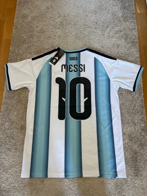 Argentina Messi matchtröja Adidas - Säljer en officiell Argentina matchtröja från Adidas med Messi och nummer 10 på ryggen. Tröjan är vit med ljusblå vertikala ränder, svarta detaljer och guldiga logotyper på bröstet. Korta ärmar och klassisk sportig passform. Perfekt för fotbollsfans!