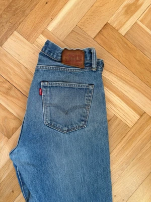 Levi's 501 blå jeans - Klassiska blå Levi's 501 jeans med rak passform och femficksdesign. Jeansens material är slitstark bomull och de har en ikonisk läderpatch bak i midjan samt den röda Levi's-taggen på bakfickan. Perfekt för dig som gillar tidlös denimstil.