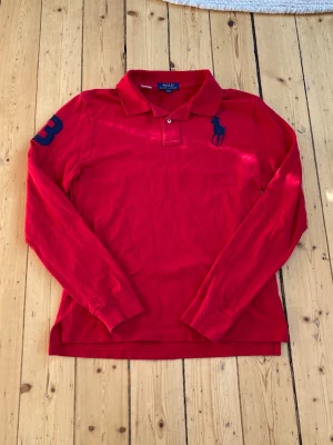 Röd långärmad piké från Polo Ralph Lauren - Snygg röd långärmad pikétröja från Polo Ralph Lauren med klassisk krage och knappar framtill. Stor blå broderad logga på bröstet och siffran 3 på ärmen. Perfekt för dig som gillar sportig och stilren look.