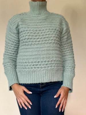 Turkos grön stickad polotröja - Mysig  stickad polotröja med strukturmönster och breda ribbade muddar vid ärmslut och nederkant. Tröjan har hög polokrage och är tillverkad i ett mjukt garn som ger en fluffig känsla. Perfekt för kyliga dagar.