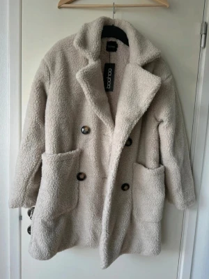 Beige teddyjacka från Boohoo - Supermjuk och oversized teddyjacka från Boohoo i beige färg. Jackan har stora fickor framtill, dubbelknäppning med knappar och bred krage. Perfekt för dig som vill ha en mysig och trendig look under kalla dagar.