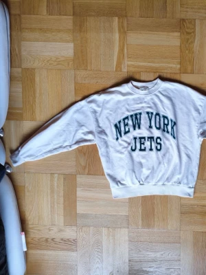Vit New York Jets hoodie - Snygg vit hoodie med trycket 'NEW YORK JETS' i grönt på bröstet. Klassisk vintage oversized modell med rund halsringning och ribbade muddar vid ärmslut och midja. Perfekt för dig som gillar sportig och avslappnad stil.