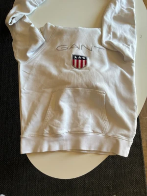 Vit hoodie från GANT med logga - Snygg vit hoodie från GANT med broderad logga och sköld med stjärnor och ränder på bröstet. Klassisk känguruficka framtill och huva. Perfekt för en avslappnad och stilren look. Tillverkad i mjukt bomullsmaterial.