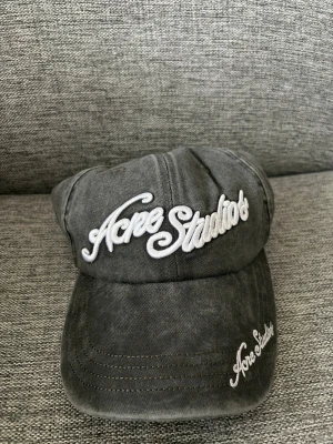 Acne studios keps - Säljer en grå keps från Acne Studios med vit broderad logga både framtill och på skärmen. Kepsen har en klassisk böjd skärm och är tillverkad i ett slitstarkt bomullstyg med tvättad look. Perfekt accessoar för en avslappnad och trendig stil.