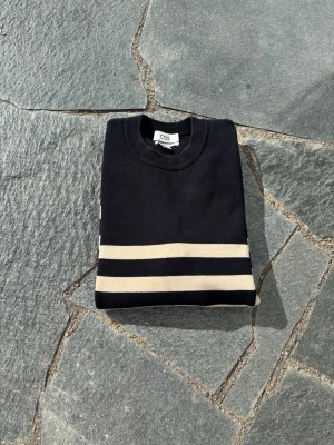 Randig stickad tröja  - Snygg stickad tröja från COS i svart med breda beigea ränder. Tröjan har rund halsringning och långa ärmar. Perfekt för dig som gillar en klassisk men modern look. Materialet känns mjukt och håller värmen bra.