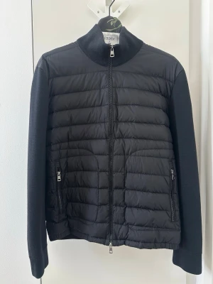 Moncler Svart jacka cardigan - Snygg svart pufferjacka från Moncler med quiltad framsida och stickade ärmar och rygg. Jackan har hög krage, två dragkedjefickor fram och Moncler-logga på ärmen. Perfekt för kalla dagar och ger en stilren look. Small stretch at bottom. Storlek xxl passar som L